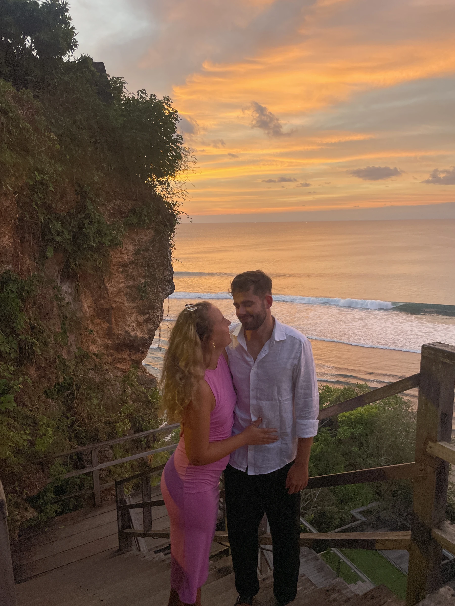 Diana und Laurin umarmen sich auf einer Holztreppe vor dem Meer im Sonnenuntergang